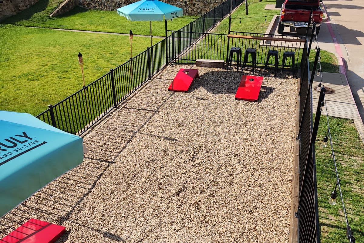 Bright red painted cornhole boards on a gravel outdoor patio beside bar seating and umbrellas