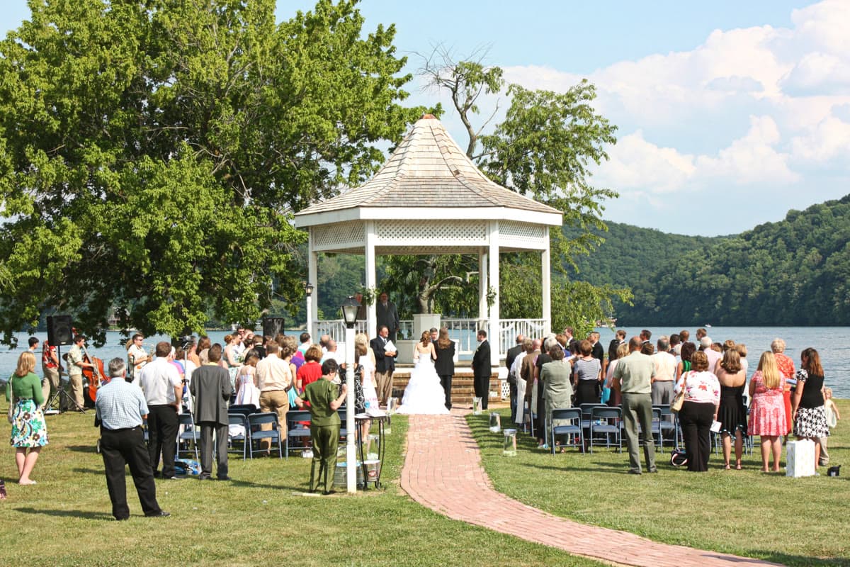 Outdoor wedding ceremony at a lakeside gazebo with guests seated on lawn chairs