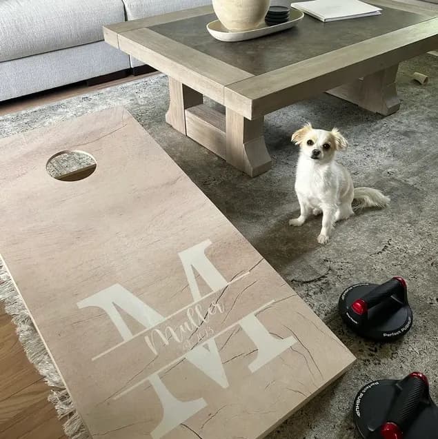 Light wood cornhole board printed with a large split M monogram and Muller in script, lying on a living-room rug next to a small dog