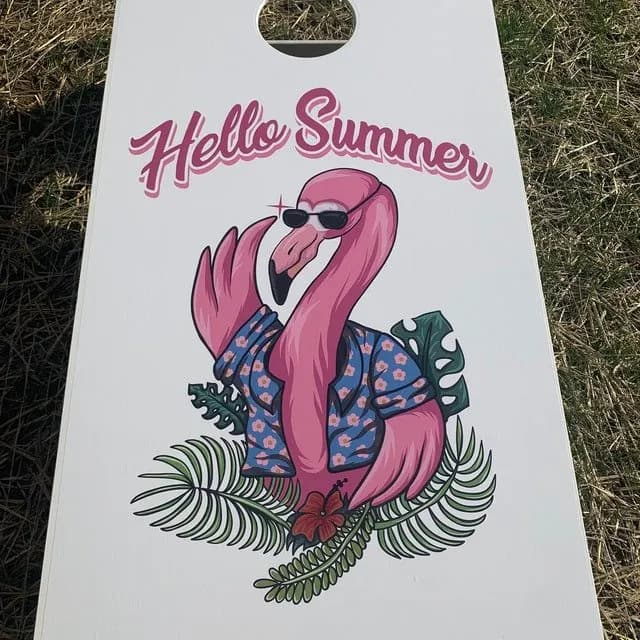 White cornhole board on grass printed with Hello Summer and a pink flamingo in a floral shirt among tropical leaves