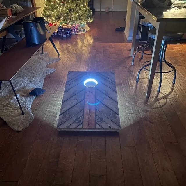 Chevron wood-plank cornhole board on a hardwood floor with a blue LED ring lighting the hole, a Christmas tree in the background
