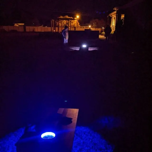 Night backyard cornhole lane with a blue LED hole in the foreground and a distant board under a small spotlight