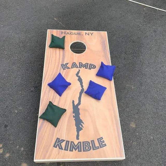 Wood-grain cornhole board printed with Hague, NY, a lake silhouette, and Kamp Kimble, with green and blue bags on the face