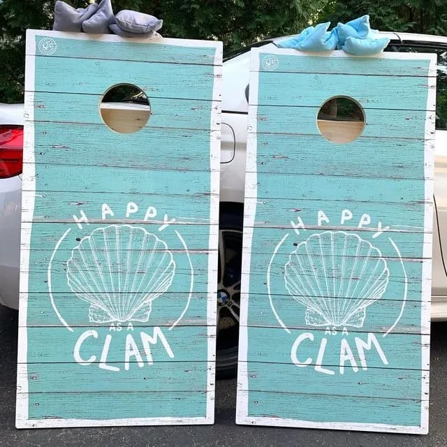 Pair of distressed teal plank cornhole boards printed with a scallop shell and Happy as a Clam, standing in front of a white car