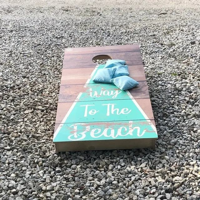 Cornhole board on gravel with a teal Way to the Beach triangle on stained wood and four light blue bags stacked on the face