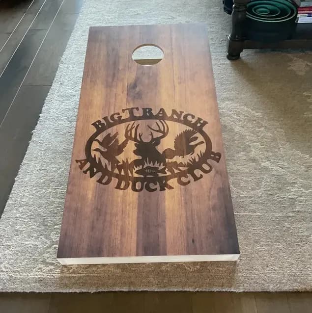Dark stained cornhole board on a rug printed with a Big T Ranch and Duck Club oval of a deer, ducks, and a turkey