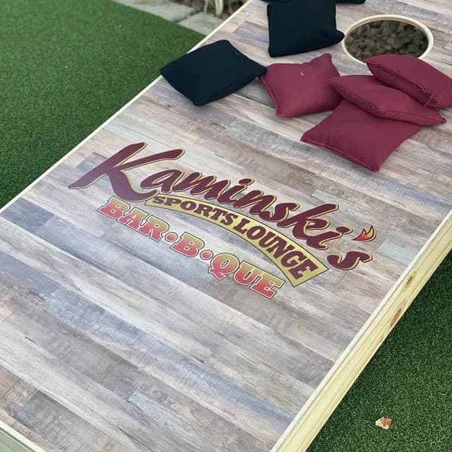 Gray wood-look cornhole board on turf printed with a Kaminski's Sports Lounge Bar-B-Que logo and black and maroon bags