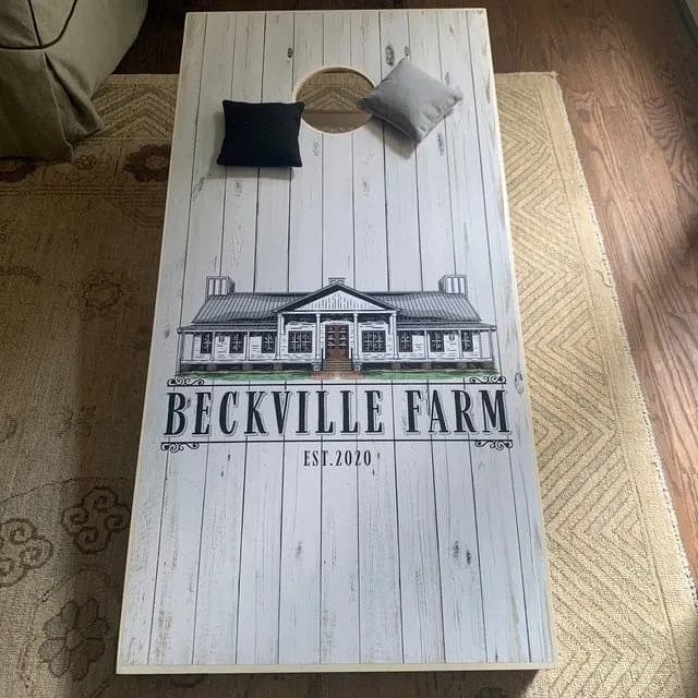 Whitewashed plank cornhole board printed with a farmhouse drawing, Beckville Farm, and Est. 2020, plus a black bag and a gray bag