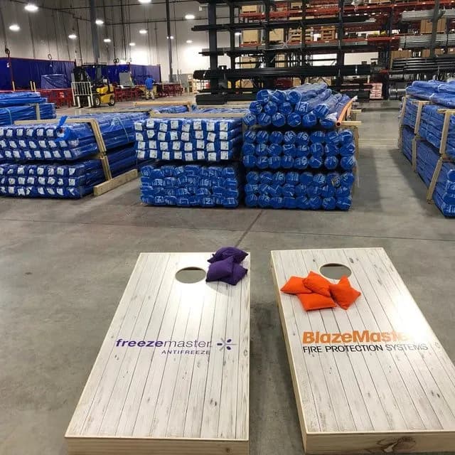 Two whitewashed cornhole boards in a warehouse printed with Freeze Master Antifreeze and BlazeMaster Fire Protection Systems logos, with purple and orange bags