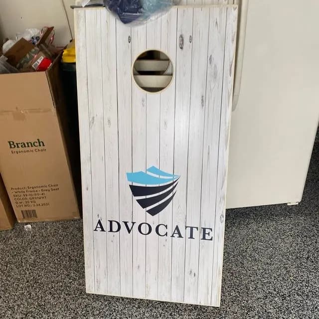 Whitewashed plank cornhole board printed with a blue shield mark and the word Advocate