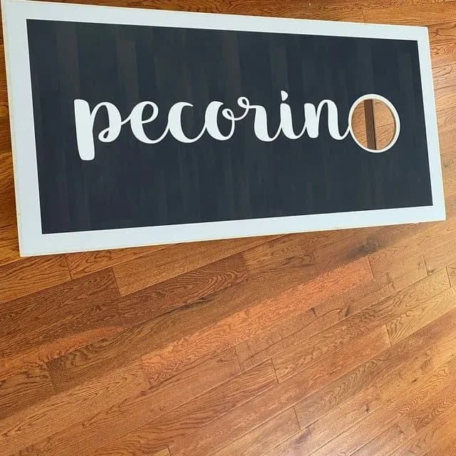 Dark cornhole board with a white frame lying on a wood floor, printed with pecorino in white script
