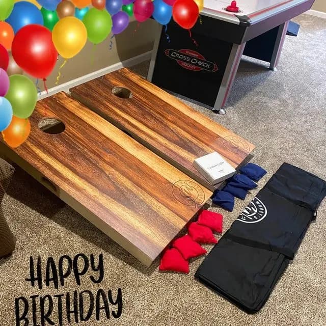 Pair of dark live-edge wood cornhole boards on a carpet with red and navy bags, a black carry case, and a birthday overlay