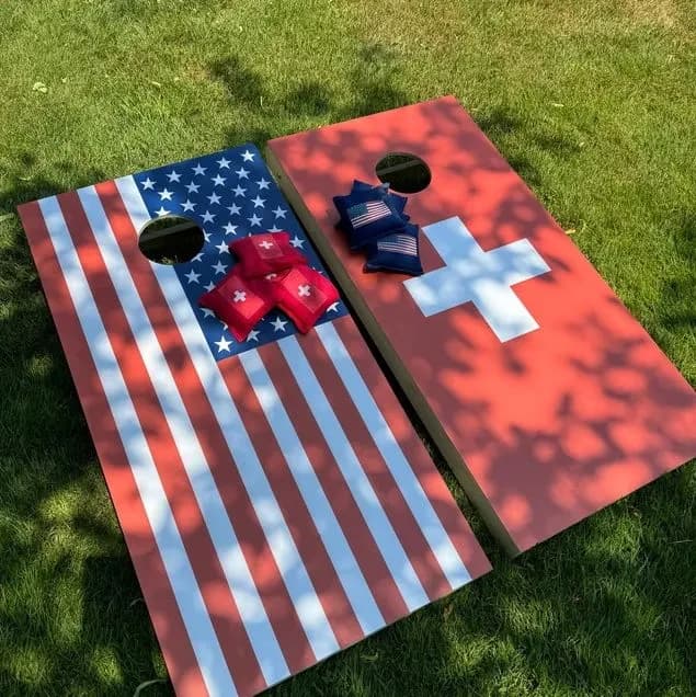 American flag cornhole board beside a red board with a white cross on a lawn, with flag-print bags and red-cross bags on the faces
