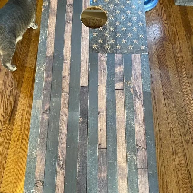 Weathered wood-plank American flag cornhole board on a hardwood floor, with a cat standing along the left edge