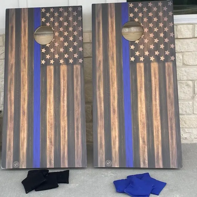 Pair of wood-flag cornhole boards with a thin blue stripe, black bags, and blue bags on a stone patio