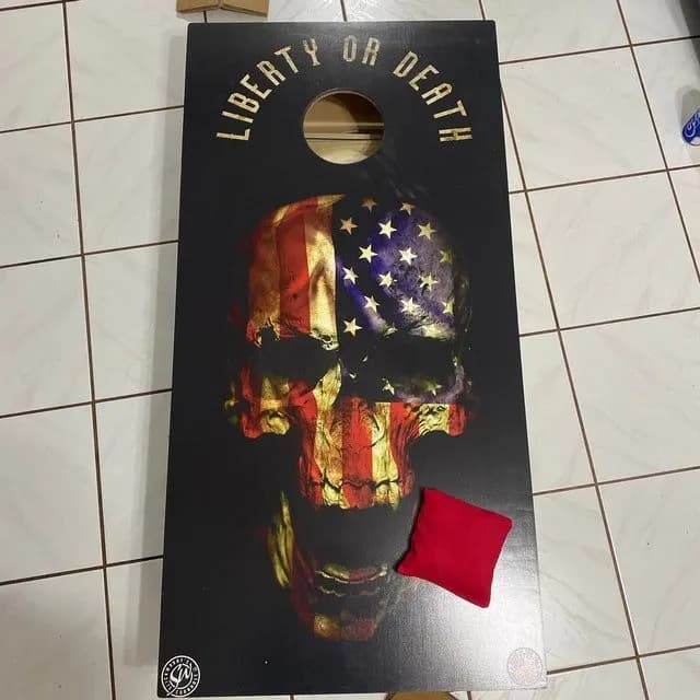 Black cornhole board printed with Liberty or Death over a skull filled with a US flag, and a red bag on the face
