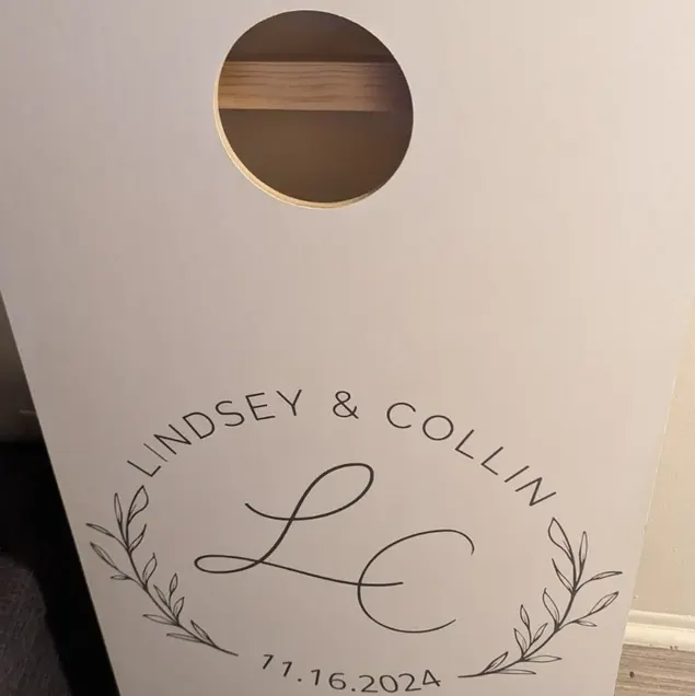 White cornhole board printed with a leaf wreath, LC initials, two names, and a November 2024 date