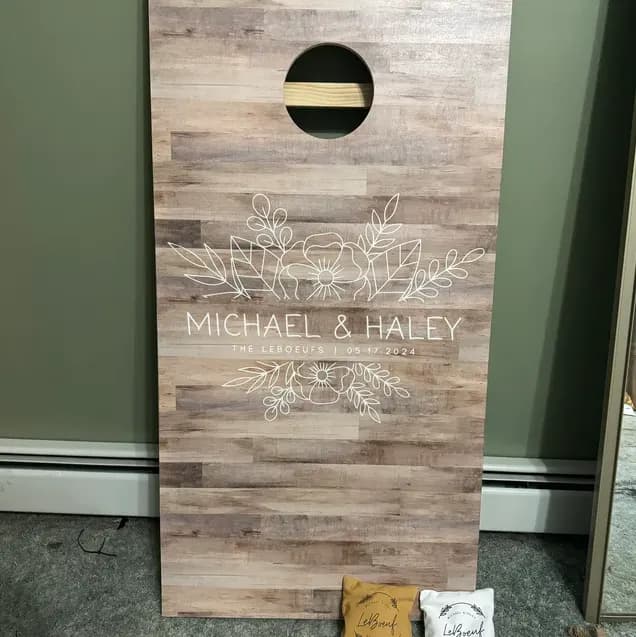 Light weathered-wood cornhole board with a white line-drawn floral, two names, a date, and gold and white bags at the bottom
