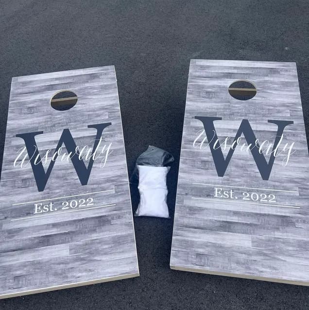 Pair of gray wood-look cornhole boards printed with a large W, a script last name, and Est. 2022, with a bag of white bags between them
