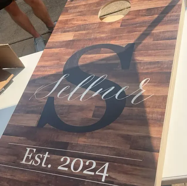 Dark wood-plank cornhole board printed with a large S, Sellner in script, and Est. 2024