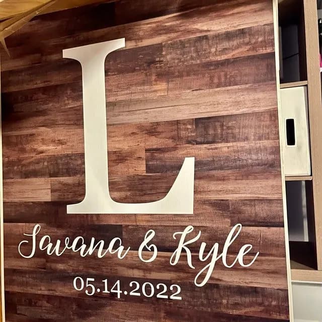 Close view of a dark wood-plank cornhole face with a large white L, two names in script, and 05.14.2022