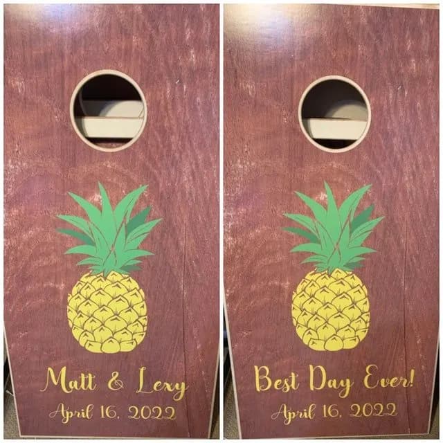 Two burgundy wood cornhole boards printed with a yellow pineapple, names or Best Day Ever, and an April 2022 date